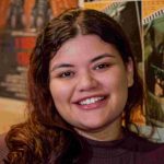 Mulher sorrindo em frente a cartazes de filmes, possivelmente em um cineclube.