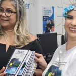 Mulheres sorrindo, segurando livros, em evento de distribuição literária para escolas.