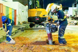 Operários trabalham em obras de saneamento em Manaus, utilizando máquinas e ferramentas.