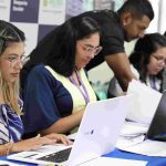 Pessoas em processo seletivo da Seduc preenchendo documentos e utilizando laptops.