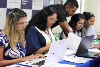 Pessoas em processo seletivo da Seduc preenchendo documentos e utilizando laptops.