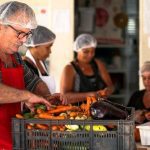 Pessoas preparando vegetais em cozinha, com foco em um homem com avental vermelho e touca.