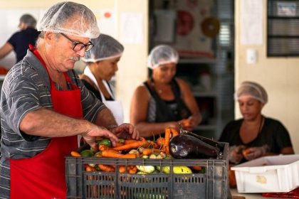 Pessoas preparando vegetais em cozinha, com foco em um homem com avental vermelho e touca.