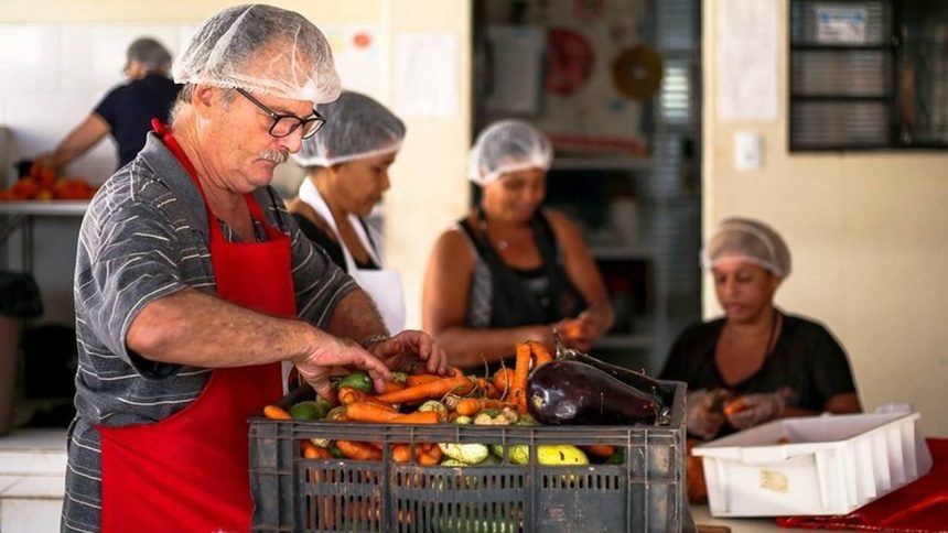 Pessoas preparando vegetais em cozinha, com foco em um homem com avental vermelho e touca.