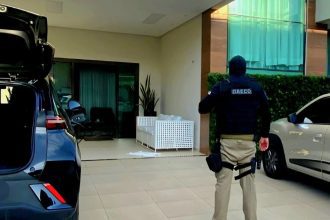 Policial em ação em frente a uma residência, possivelmente em operação contra crime organizado.