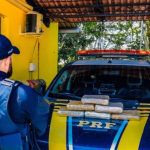 PRF apreende drogas em operação na BR-319. Policial em frente à viatura com drogas apreendidas.