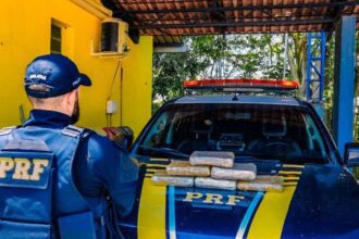 PRF apreende drogas em operação na BR-319. Policial em frente à viatura com drogas apreendidas.