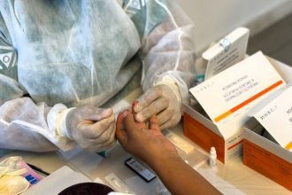 Profissional de saúde realizando teste rápido em paciente, usando luvas e jaleco.