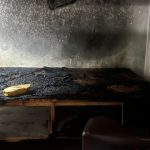 Quarto destruído por incêndio, com cama carbonizada e paredes enegrecidas.