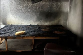 Quarto destruído por incêndio, com cama carbonizada e paredes enegrecidas.