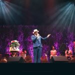 Show do cantor Antônio Bahia no Teatro ICBEU, com banda e cenário com flores.