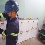 Trabalhadores instalando azulejos em um banheiro, usando capacetes de segurança e ferramentas.