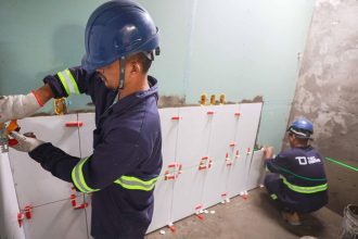 Trabalhadores instalando azulejos em um banheiro, usando capacetes de segurança e ferramentas.