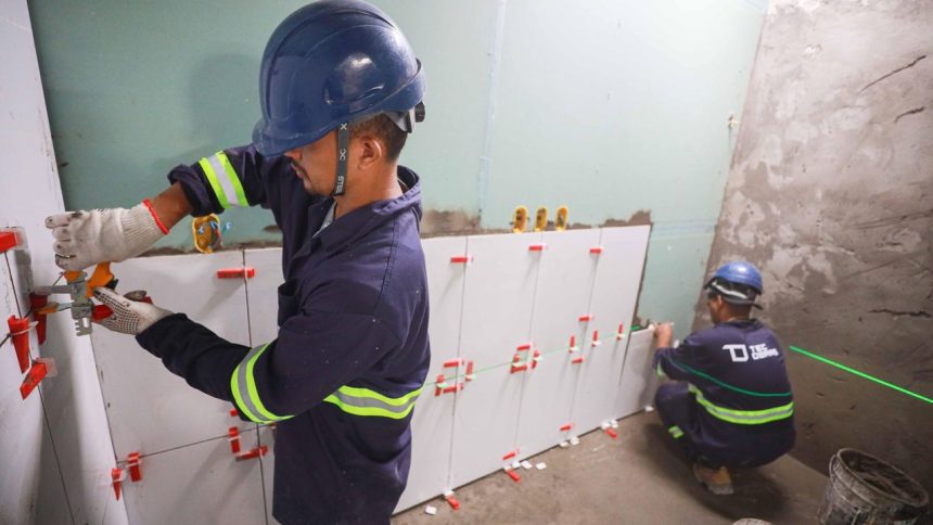 Trabalhadores instalando azulejos em um banheiro, usando capacetes de segurança e ferramentas.