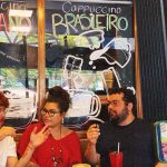 Ateliê 23 Três pessoas em um café com um letreiro de 'Cappuccino' em português e italiano, e uma tela de celular gravando.