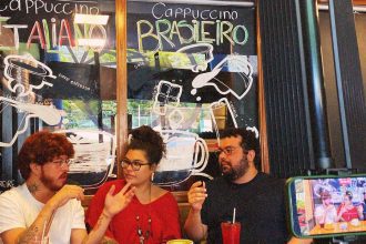 Ateliê 23 Três pessoas em um café com um letreiro de 'Cappuccino' em português e italiano, e uma tela de celular gravando.