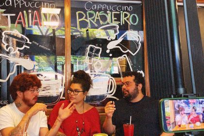 Ateliê 23 Três pessoas em um café com um letreiro de 'Cappuccino' em português e italiano, e uma tela de celular gravando.