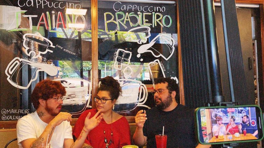 Ateliê 23 Três pessoas em um café com um letreiro de 'Cappuccino' em português e italiano, e uma tela de celular gravando.