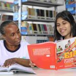Alunas estudando em biblioteca, focando em um livro sobre educação de jovens e adultos.