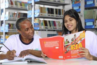 Alunas estudando em biblioteca, focando em um livro sobre educação de jovens e adultos.