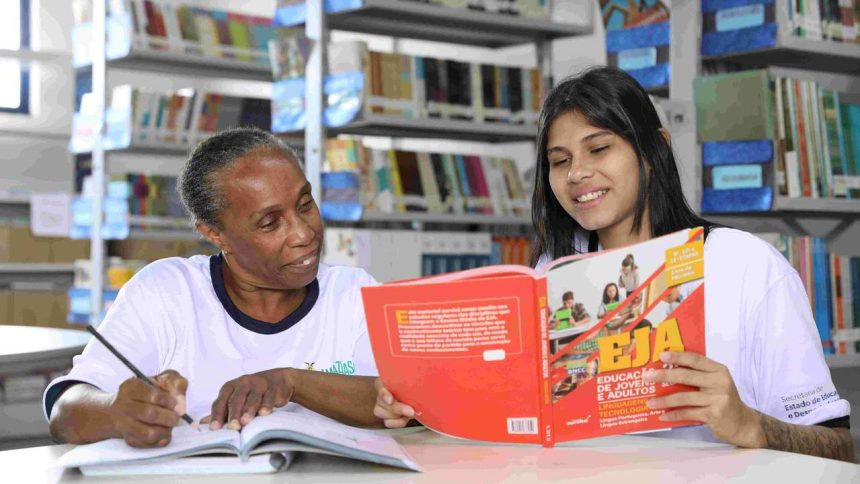 Alunas estudando em biblioteca, focando em um livro sobre educação de jovens e adultos.