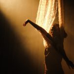 Ator em performance de dança no espetáculo 'Gaia', com luz dramática e cortina.