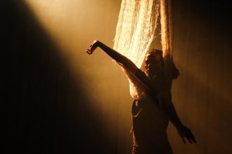 Ator em performance de dança no espetáculo 'Gaia', com luz dramática e cortina.