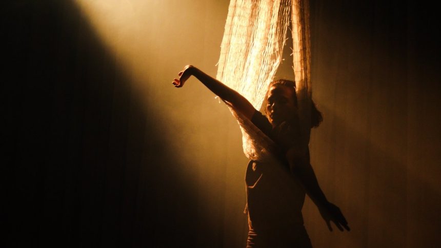 Ator em performance de dança no espetáculo 'Gaia', com luz dramática e cortina.