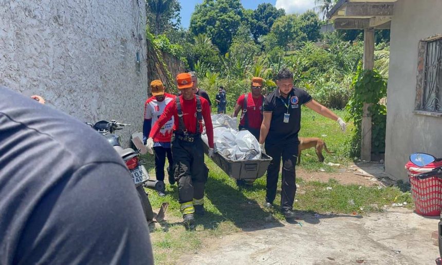 Bombeiros carregam corpo em maca em área de mata em comunidade.