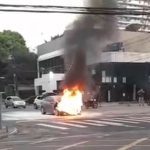Carro em chamas em cruzamento, com fumaça intensa subindo.