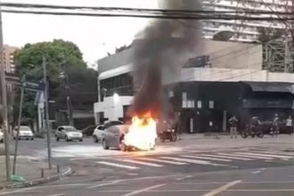 Carro em chamas em cruzamento, com fumaça intensa subindo.