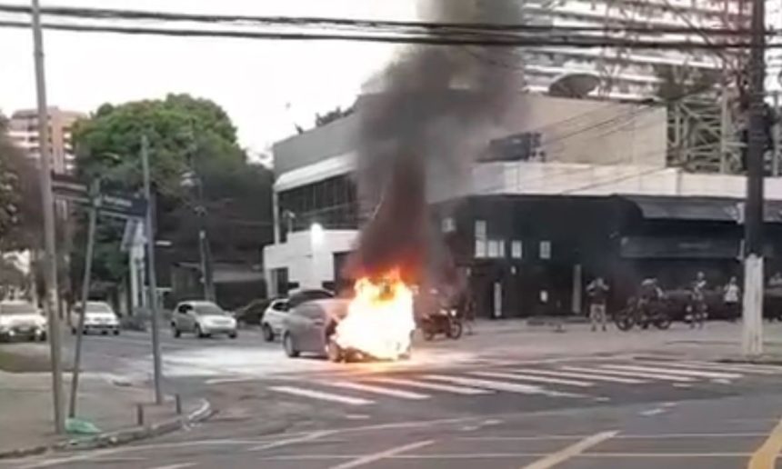 Carro em chamas em cruzamento, com fumaça intensa subindo.