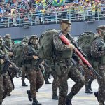 Desfile militar com soldados marchando, equipados com uniformes camuflados e mochilas.