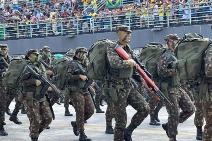Desfile militar com soldados marchando, equipados com uniformes camuflados e mochilas.