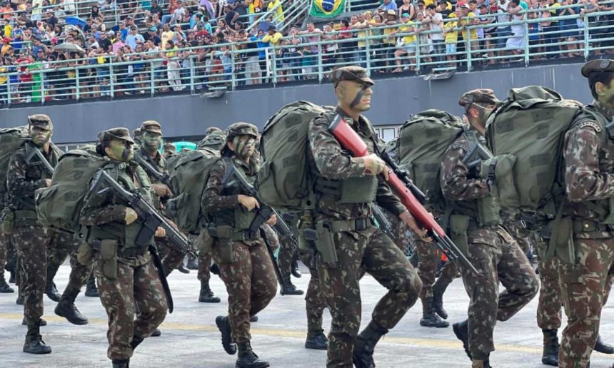 Desfile militar com soldados marchando, equipados com uniformes camuflados e mochilas.