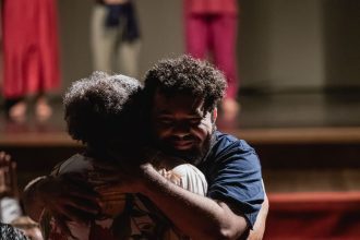 Dois homens se abraçam no palco, com outros atores ao fundo, em evento do Ateliê 23.