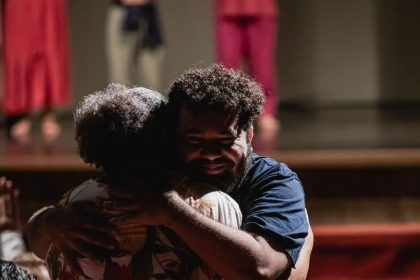Dois homens se abraçam no palco, com outros atores ao fundo, em evento do Ateliê 23.