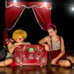 Dois palhaços sentados no chão em um palco de circo, com uma mala vermelha escrita "Circo".