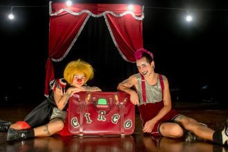 Dois palhaços sentados no chão em um palco de circo, com uma mala vermelha escrita "Circo".