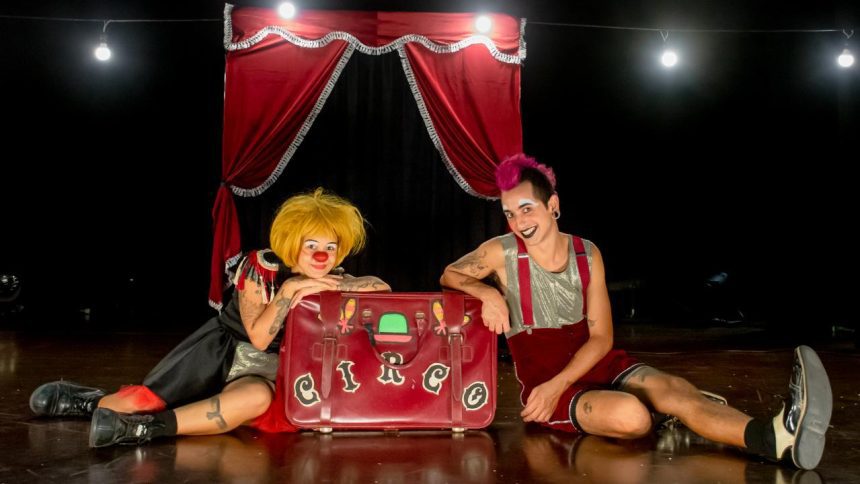 Dois palhaços sentados no chão em um palco de circo, com uma mala vermelha escrita "Circo".
