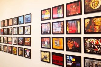 Exposição de arte com várias molduras pretas penduradas em uma parede branca, com obras de arte variadas.