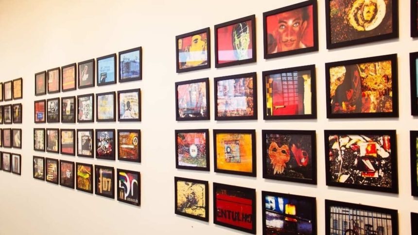 Exposição de arte com várias molduras pretas penduradas em uma parede branca, com obras de arte variadas.