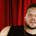 Flávio Andradde em close com expressão de surpresa, apontando para cima, em palco de teatro.