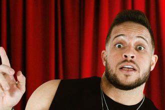 Flávio Andradde em close com expressão de surpresa, apontando para cima, em palco de teatro.