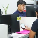 Funcionários em escritório, trabalhando em computadores e verificando um smartphone. Cena de escritório.