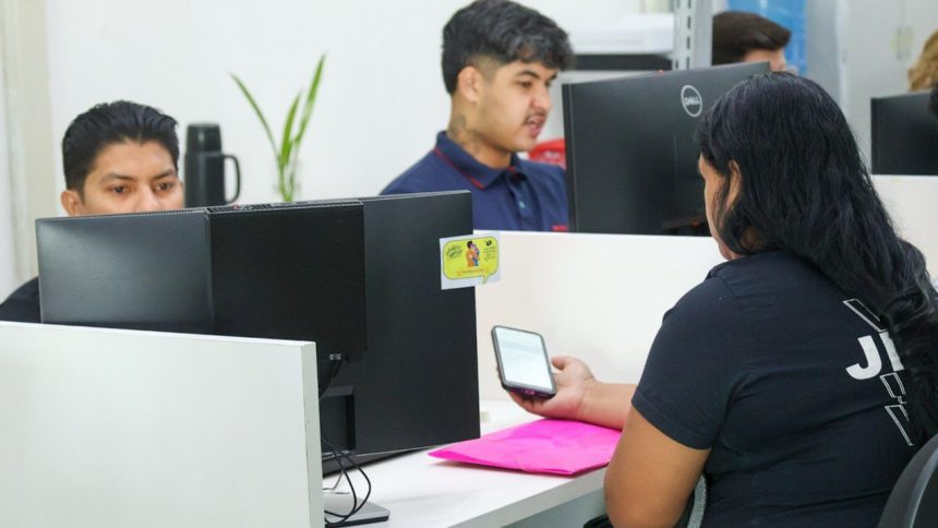 Funcionários em escritório, trabalhando em computadores e verificando um smartphone. Cena de escritório.