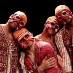 Grupo de dançarinos com máscaras coloridas e roupas tradicionais se apresenta no palco.