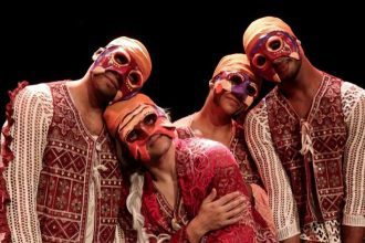 Grupo de dançarinos com máscaras coloridas e roupas tradicionais se apresenta no palco.
