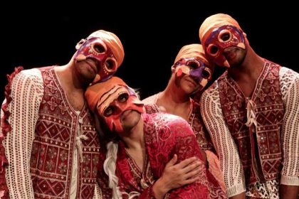 Grupo de dançarinos com máscaras coloridas e roupas tradicionais se apresenta no palco.
