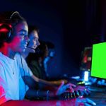 Grupo de jovens gamers com fones de ouvido, jogando em computadores com telas verdes.